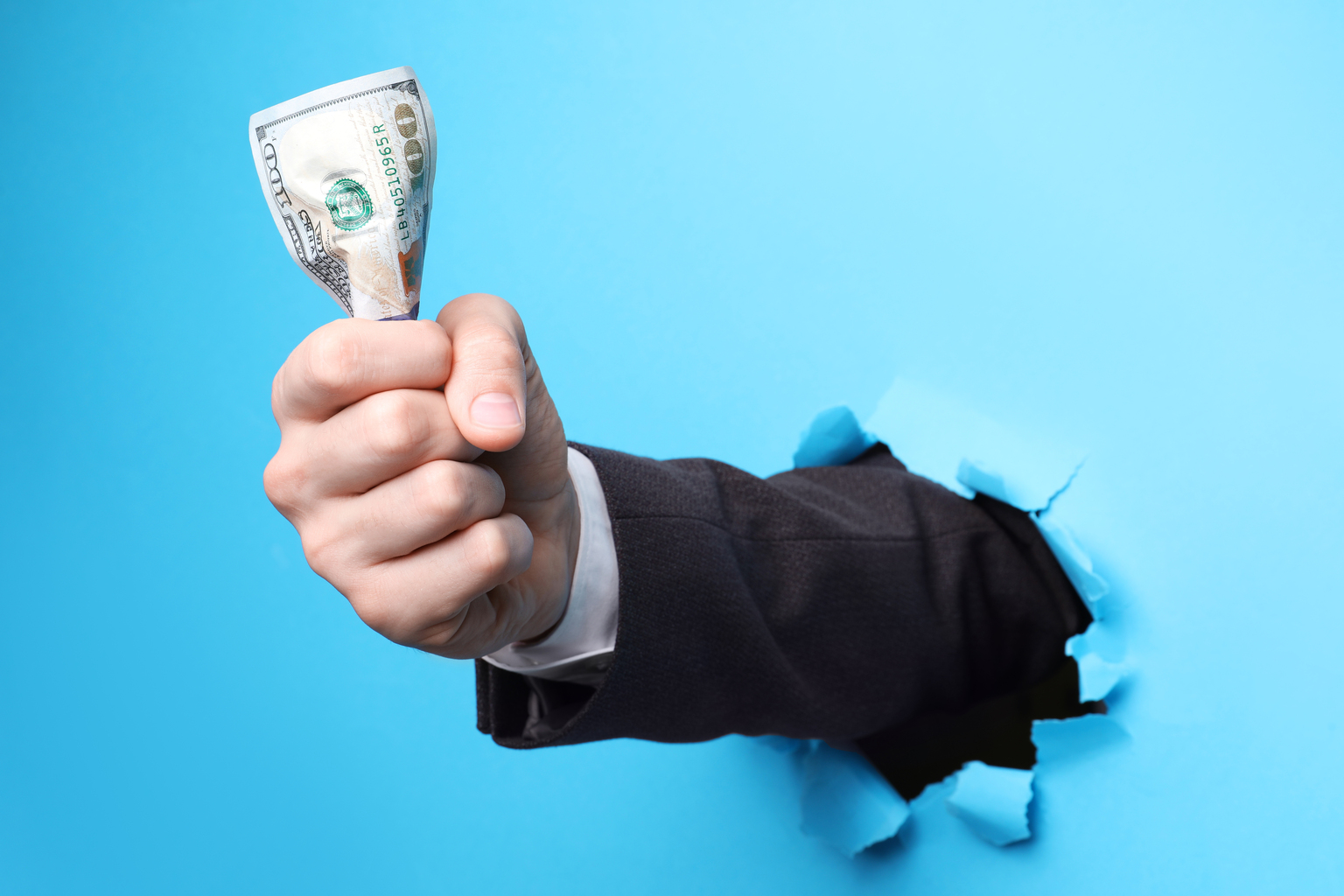 Businessman breaking through light blue paper with money in fist, closeup