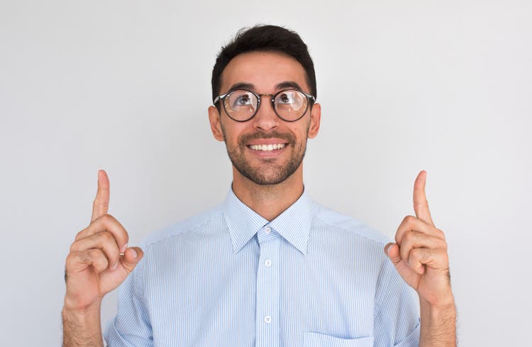 Horizontal closeup shot of happy satisfied Caucasian male points upwards with both index fingers, notices something amazing above head, looikng up, wearing round spectacles, against white studio wall