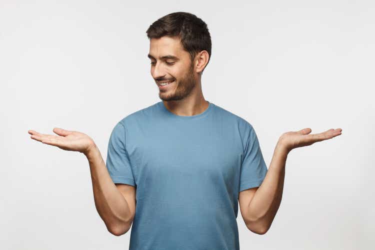 Choice concept. Portrait of young man choosing between 2 different options, holding two hands with empty space, isolated on gray background