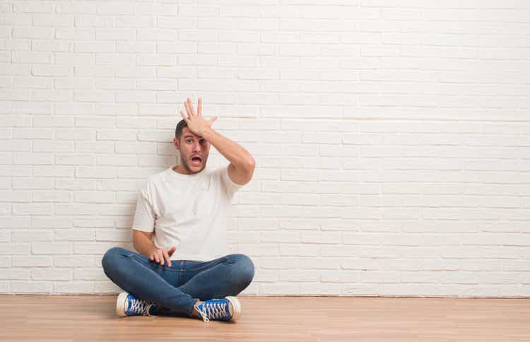 Young caucasian man sitting on the floor over white brick wall surprised with hand on head for mistake, remember error. Forgot, bad memory concept.