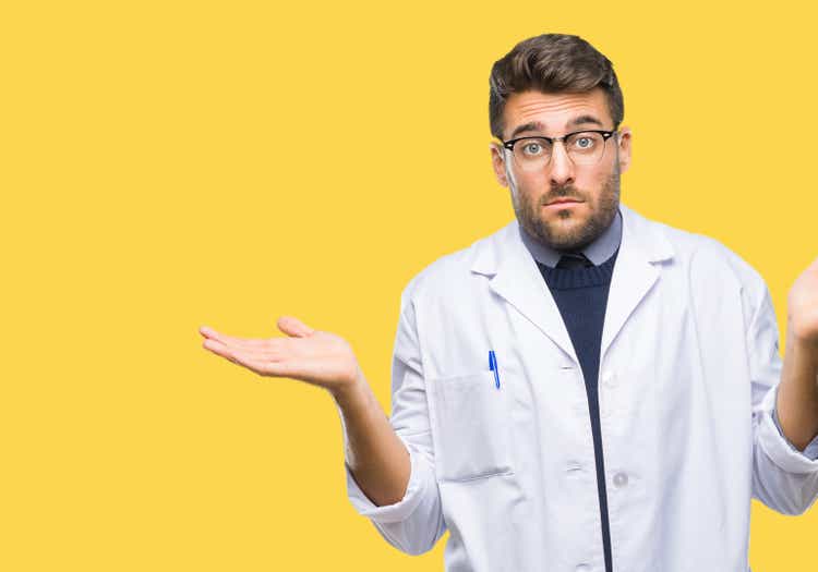 Young handsome man wearing doctor, scientis coat over isolated background clueless and confused expression with arms and hands raised. Doubt concept.