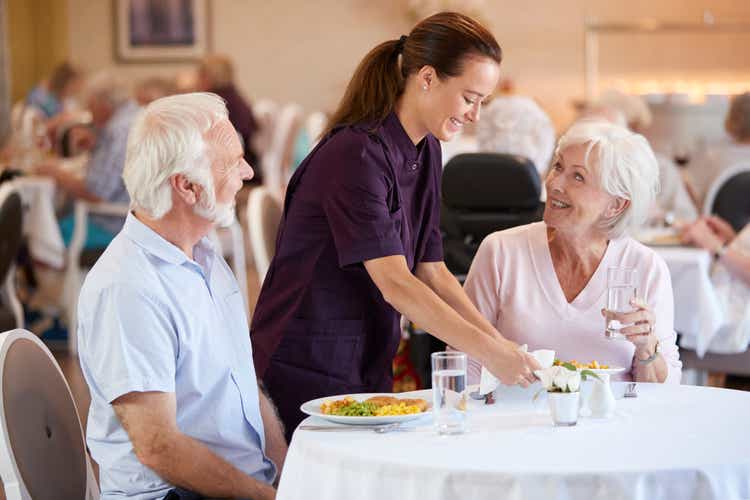 Senior Couple Being Served With Meal By Carer In Dining Room Of Retirement Home