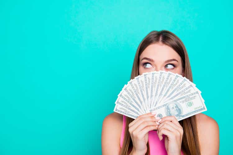 Attractive gorgeous young amazed girl wearing pink blouse, excited, covering face with dollar banknotes. Copy space. Isolated over bright vivid blue teal, turquoise background