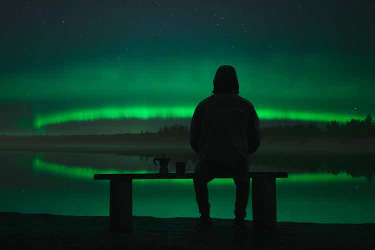 A man sits on a bench near the lake and looks at the northern lights. Coffee maker and mug nearby on the bench.