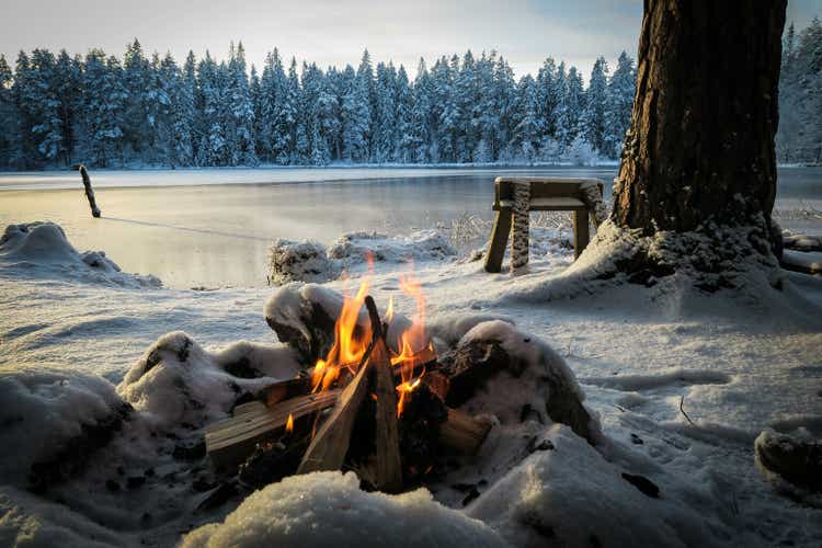 En lägereld i förgrunden en solig vinterdag vid en frusen skogssjö i ett snöigt vinterlandskap utan människor