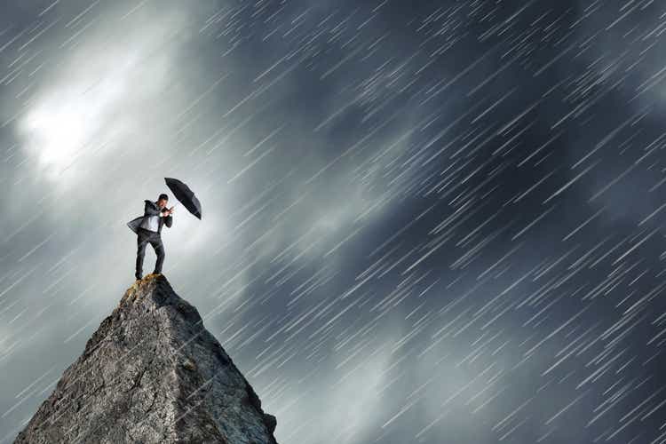 Businessman Holding Umbrella Stands On Mountain Peak In Rainstorm