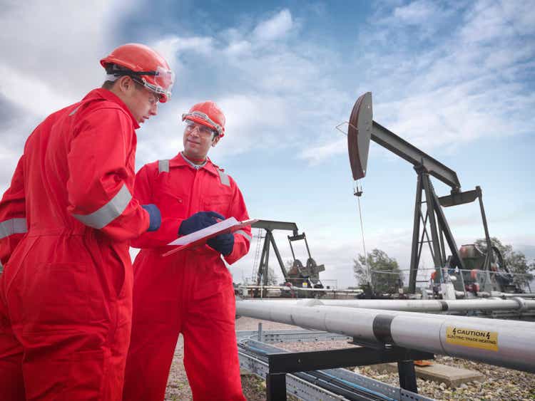 Two workers looking at a clipboard in the foreground with onshore oil pumps above oil wells (nodding donkeys/pumpjacks) in the background