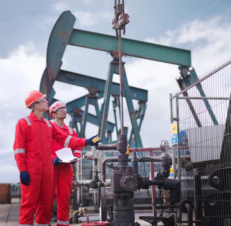 Two workers taking a reading and looking up, next to oil pumps above onshore oil wells (nodding donkeys/pumpjacks)