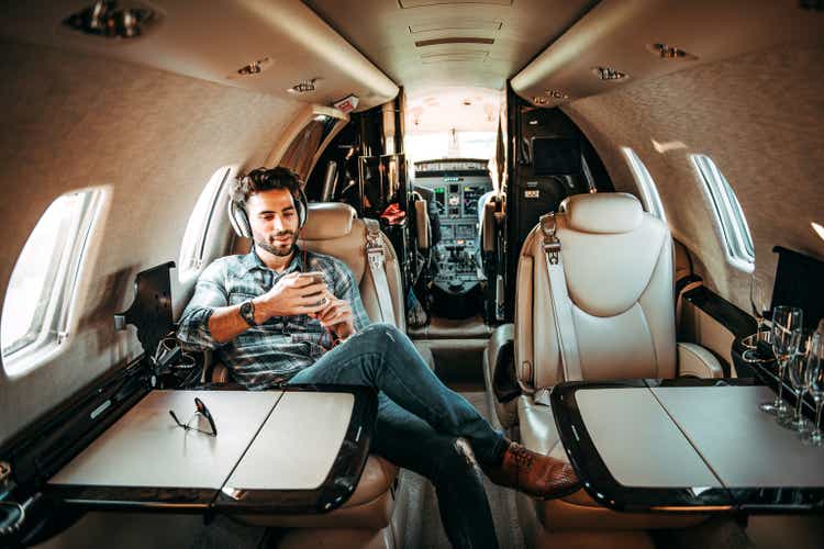Young rich man listening to music over the headphones and using a mobile phone while sitting in a private jet