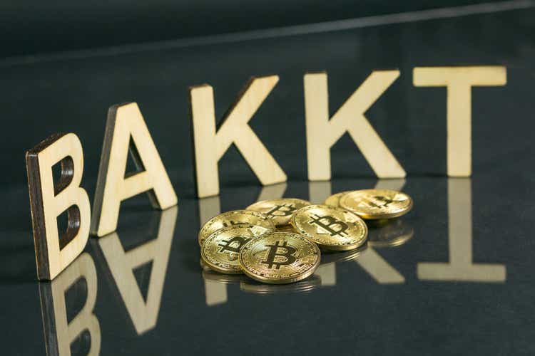 Bitcoin coins in front of bakkt sign made of wood with reflection on the table, Slovenia - December 27th