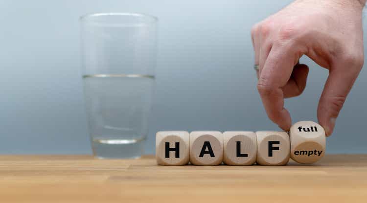 Hand turns a dice and changes the expression "half empty" to "half full". A half full glass of water is standing in front of a grey background.
