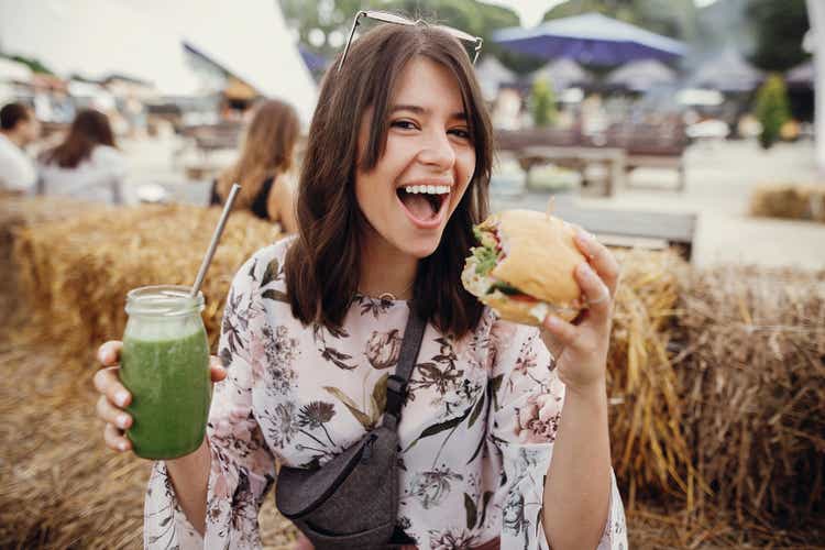 Stylish hipster girl in sunglasses holding delicious vegan burger and smoothie in glass jar in hands at street food festival. Happy boho woman holding burger and drink in summer street