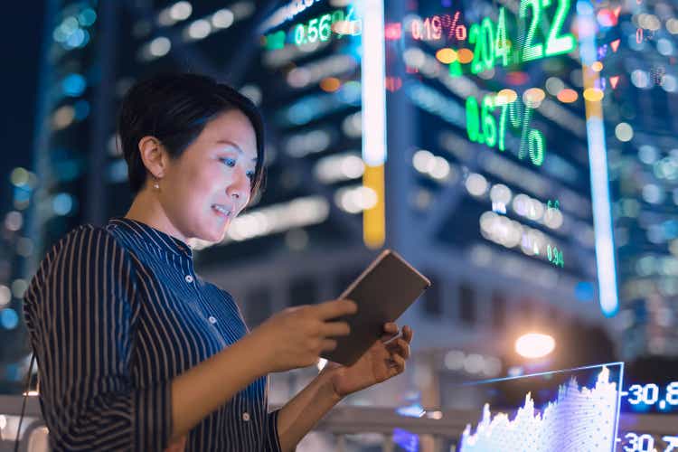 Asian businesswomen checking stock market data on tablet before Hong Kong financial display board