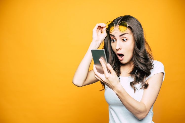 Oh, my! Gorgeous girl with a phone in her hand is surprised by something she saw on the screen is holding her glasses in front of bright yellow background.