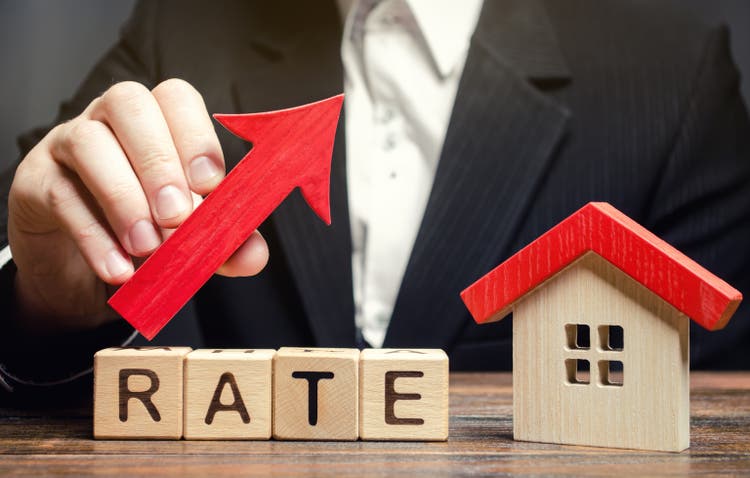 A man holds a red arrow up above the word Rate and a wooden house. The concept of raising interest rates on mortgages. The increase in property tax rates. Real estate capitalization. Insurance.