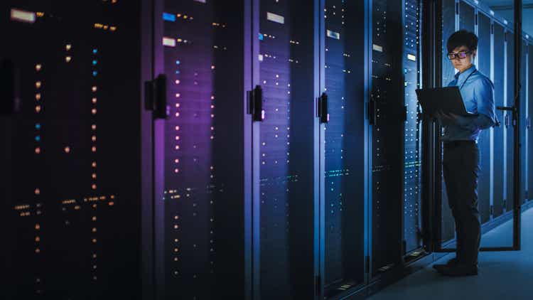 In Dark Data Center: Male IT Specialist Stands Beside the Row of Operational Server Racks, Uses Laptop for Maintenance. Concept for Cloud Computing, Artificial Intelligence, Supercomputer, Cybersecurity. Neon Lights