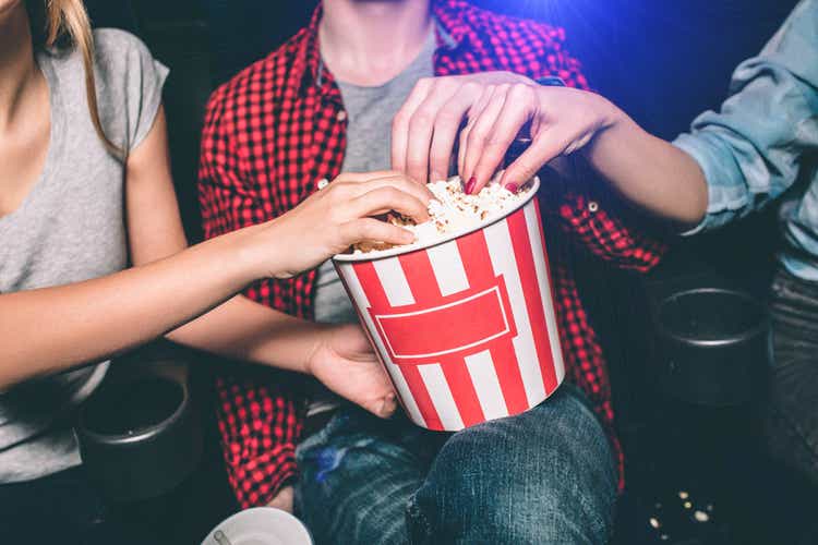 Close up of red with white basket of popcorn that both girl and guy are holding. All of them are taking popcorn out of basket. Сut view.