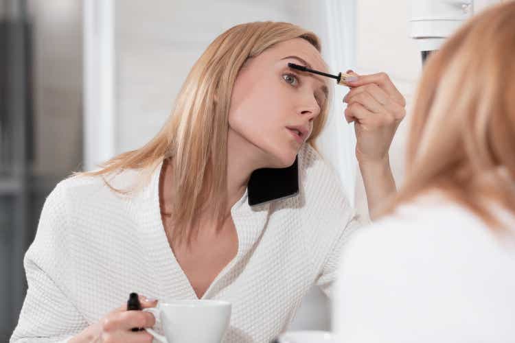 Aantrekkelijke blonde vrouw in witte badjas staat in de badkamer per spiegel. Past make-up, spreekt op mobiele telefoon en drinkt koffie. "S ochtends vergoedingen, geen tijd, doen veel actie op hetzelfde moment.