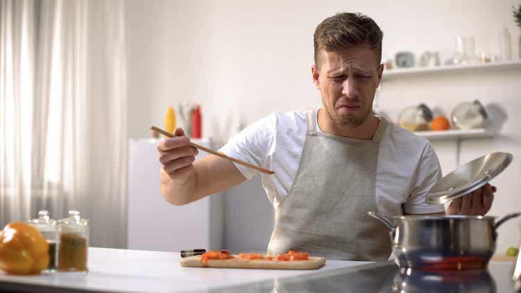 Young man tasting cooked food with disgusted face expression, funny grimacing