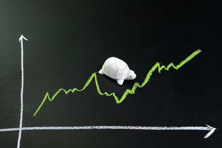 Slow but stable investment or low fluctuate stock market concept, miniature figure turtle or tortoise walking on chalkboard with drawing green price line graph of stock market value