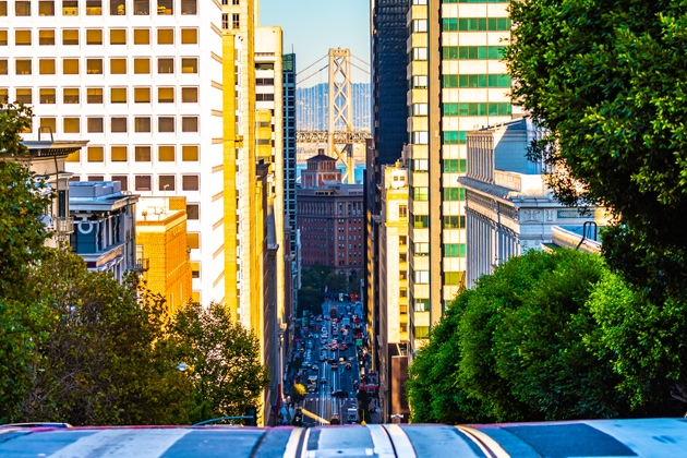 City street view in San Francisco, California uphill looking down between buildings and other architecture at Oakland Bay Bridge over the ocean.