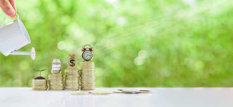 Long-tern investment, time value of money concept : Clock, US dollar bag, glass jar, a small tree on steps of rising coins. Hand pour water from a watering can, depicts asset growth from investment