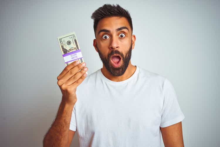 Young indian man holding dollars standing over isolated white background scared in shock with a surprise face, afraid and excited with fear expression