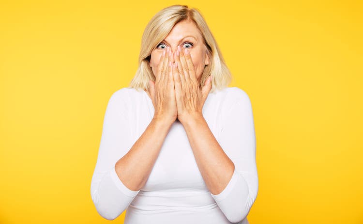 Don"t talk! Blonde Senior woman covers her mouth with help of hands and looks on camera with shock in eyes.