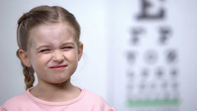 Small girl frowning, trying to see letter on vision testing table blurred vision