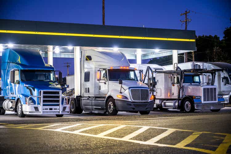 Different make and models big rig semi trucks with semi trailers standing on the truck stop parking lot under the lighted shelter in night