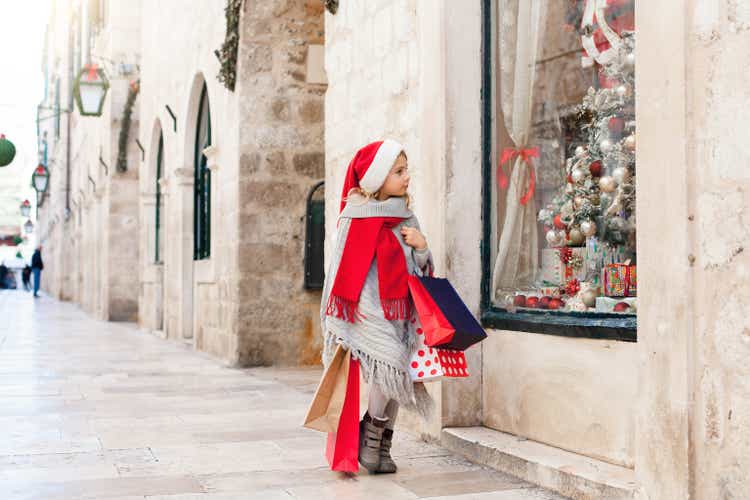 Kid with shopping bags on market street outside. Funny child is looking at store window decorated with gifts, christmas tree. Girl in red santa hat in old town. Cozy festive and New Year atmosphere.