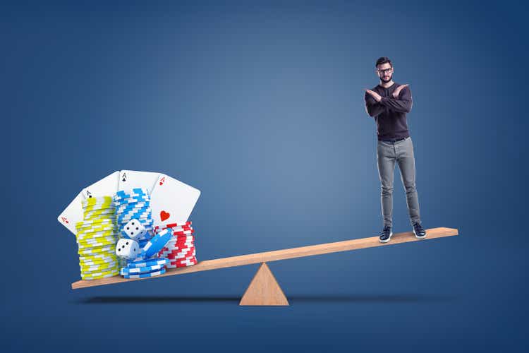 A man stands making a cross gesture with his arms while standing on a seesaw with heavier casino chips on another side.