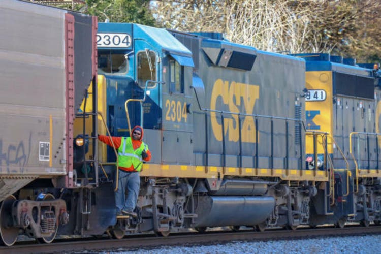 A railroad employee holding a walkie-talkie leans out from a CSX railroad locomotive,