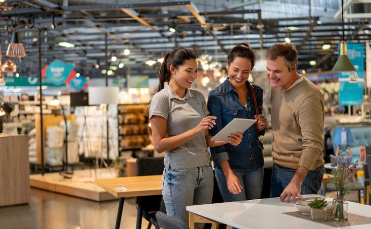Cheerful sales woman showing a design on tablet to mid adult couple looking for furniture at a home store