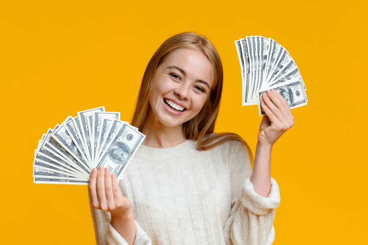 Happy millennial girl holding lots of dollars on orange background