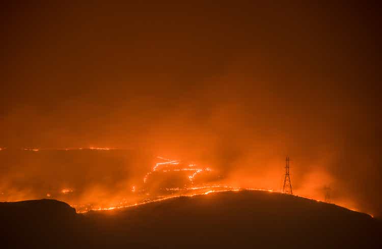 A massive wildfire burns its way in to Grand Coulee Washington at night. The fire and the city glow in the smoke night air. Silhouettes of the power line towers that connect the Grand Coulee Dam can be clearly seen.