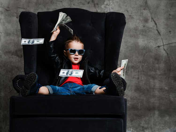 Rich kid boy in sunglasses and leather jacket is sitting in big armchair with a bundle of dollars cash, throwing money