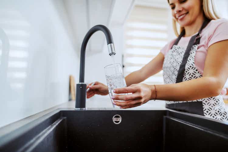 Charming smiling Caucasian blond housewife standing in kitchen and pouring water from sink. Home interior.