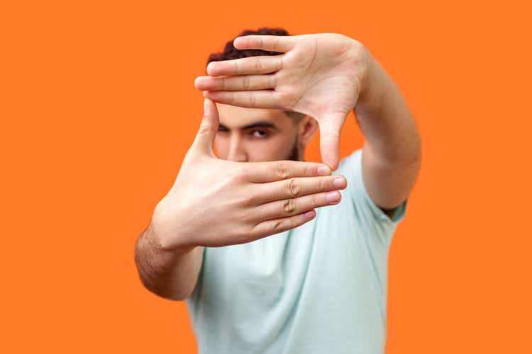 Portrait of attentive curious brunette man focusing through photo frame made of fingers. indoor studio shot isolated on orange background