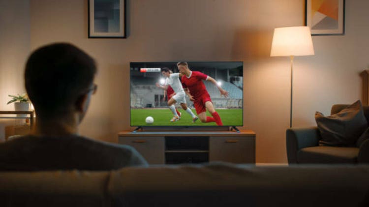Young Man in Glasses is Sitting on a Sofa and Watching TV with a Soccer Match. It"s Evening and Room at Home Has Working Lamps.