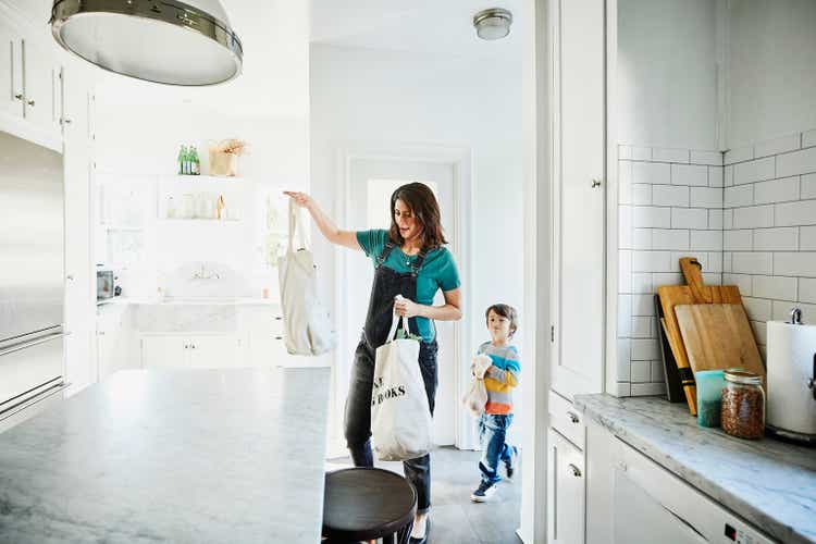 A pregnant mother carries groceries in a canvas bag to the kitchen with her little son