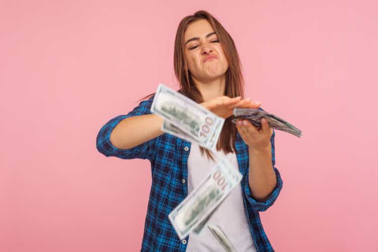 Portrait of wasteful rich girl in checkered shirt scattering dollars with arrogant grimace, boasting wealthy life