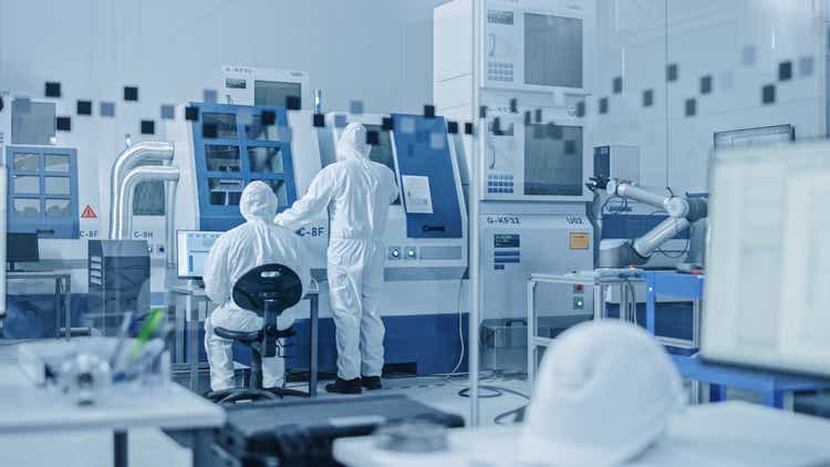 Sterile Modern Factory: Professionals in Coveralls, Masks Working on CNC Machinery. Medical Electronics Manufacturing Laboratory with High Tech Robot Arm Production Line and Contemporary Equipment