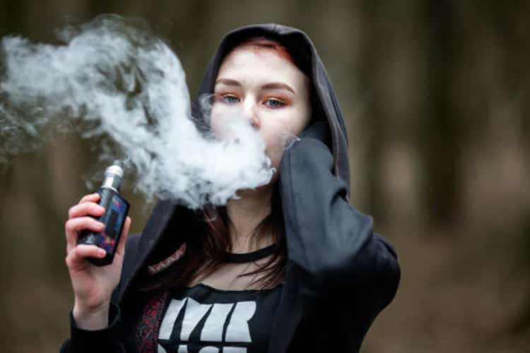 Vape teenager. Young pretty caucasian brunette girl with problem skin in the hood smoking an electronic cigarette on the street in the spring. Bad habit.
