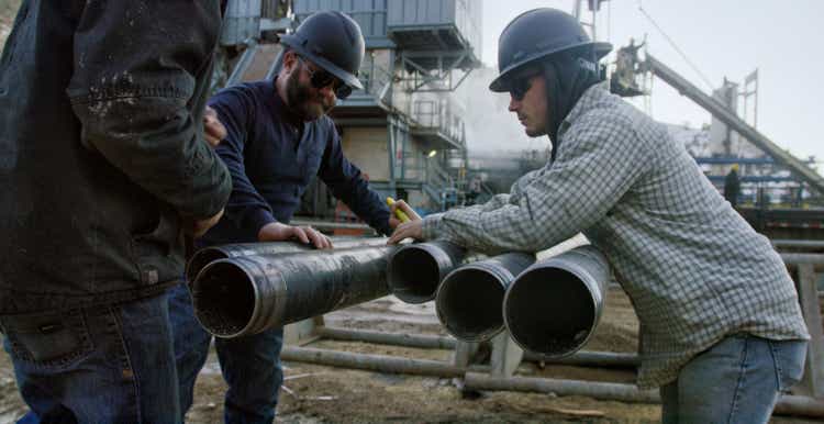 Three Oilfield Workers Remove the Drilling Pipe Thread Protector Caps at an Oil and Gas Drilling Pad Site on a Sunny Day