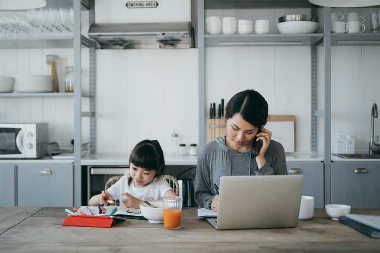 Young Asian mother working from home on a laptop and talking on the phone while little daughter is studying from home. She is attending online school classes with a digital tablet and doing homework at home