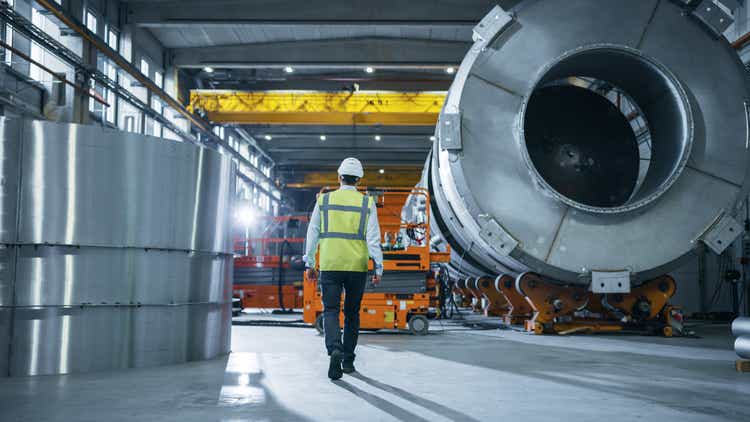 Following Shot of Heavy Industry Engineers Walking Through Manufacturing Factory. In the Background Professionals Working on Construction of Oil, Gas and Fuel Pipeline Transportation Products