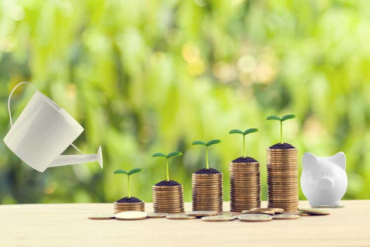 Saving money for future growth concept: Water being poured on green sprout on rows of increasing coins on wood table in the natural green background. Banking and finance, Depicts asset security for sustainable growth.