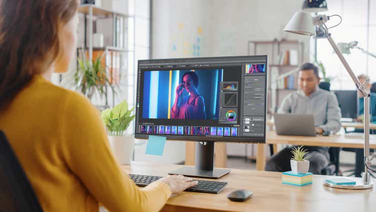 Over the Shoulder: Creative Young Woman Digital Editor Works in Photo Editing Software on Her Personal Computer with Big Display. In the Background Loft Office with Diverse Team of Young Professionals