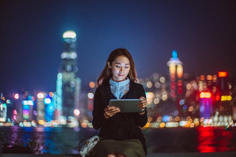 Young cheerful Asian woman using digital tablet at promenade against illuminated cityscape at night